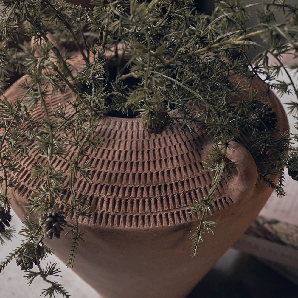 Westbrook ceramic vase in warm terracotta tones holds trailing green foliage, showing its small loop handles, tapered rounded body and rows of textured vertical marks.