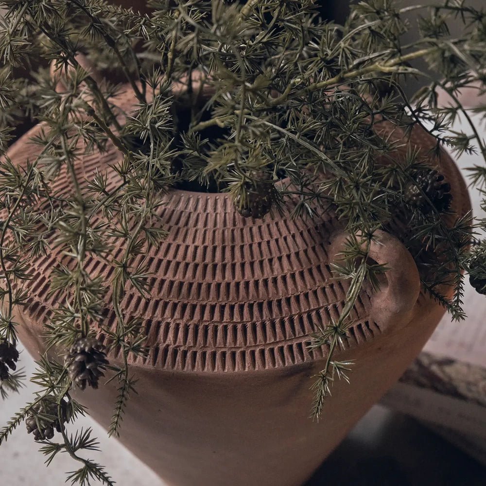 Westbrook ceramic vase in warm terracotta tones holds trailing green foliage, showing its small loop handles, tapered rounded body and rows of textured vertical marks.