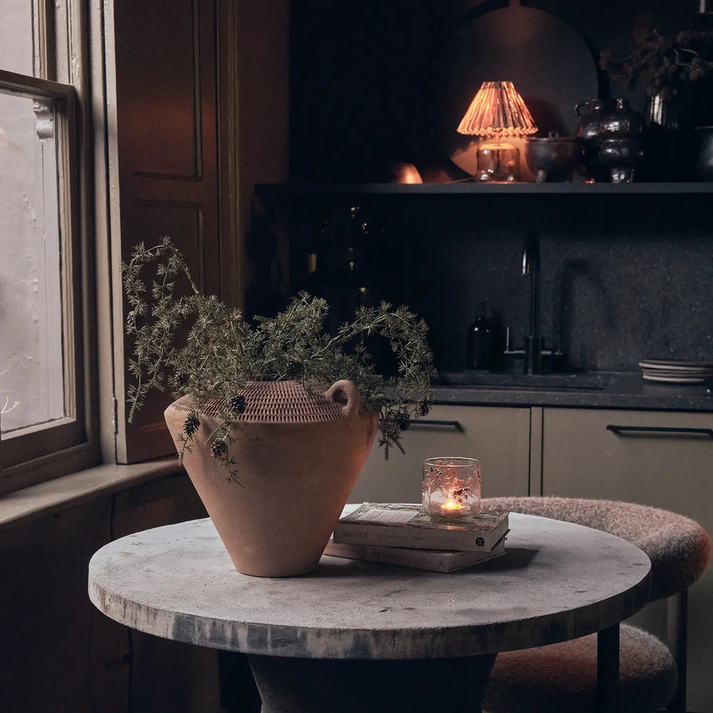 Westbrook ceramic vase with a tapered terracotta body and small loop handles holds green foliage on a round table beside a candle in a kitchen.