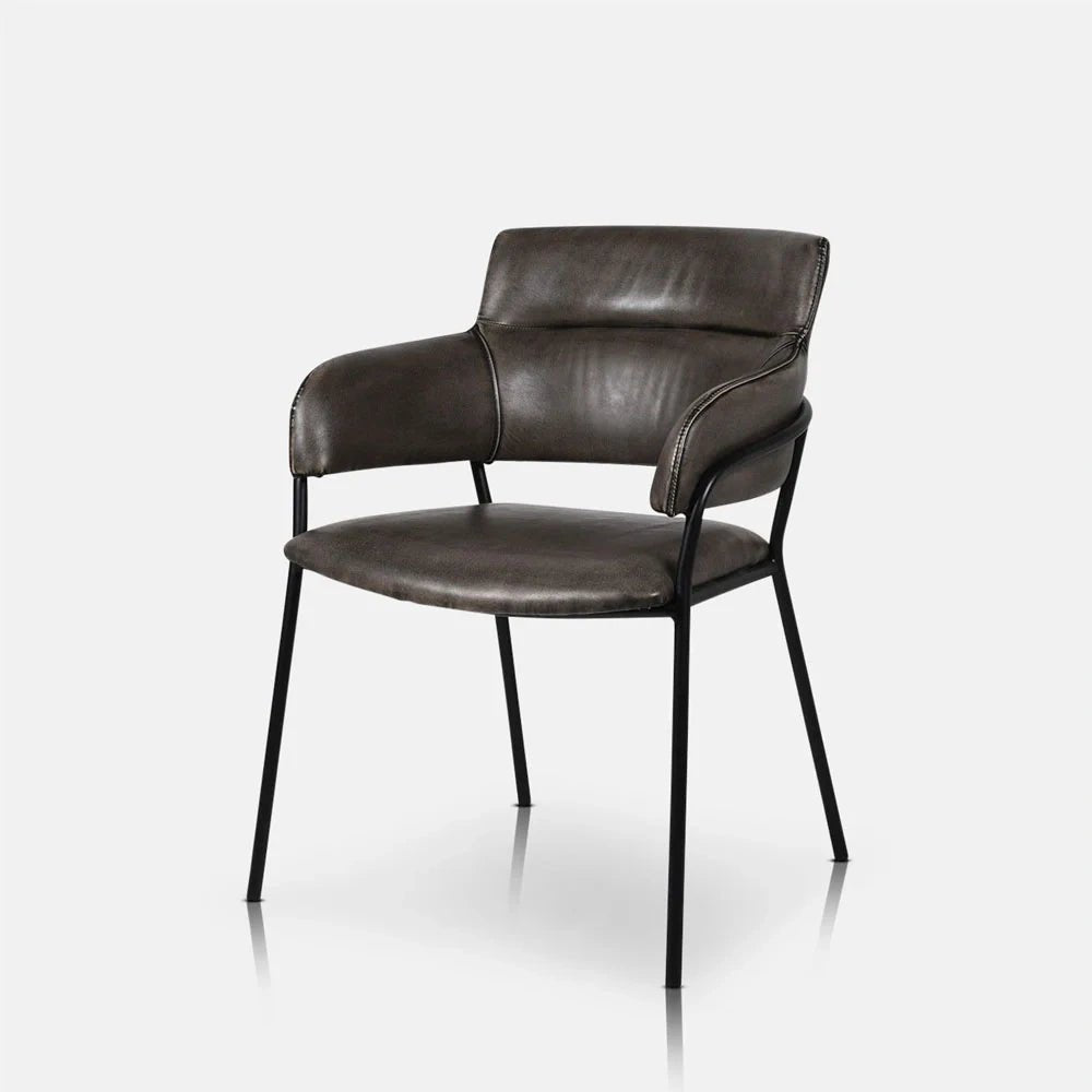 Whitcomb faux leather dining chair viewed at an angle, showing dark padded upholstery, a curved back and arms, rounded seat and slender black metal legs.