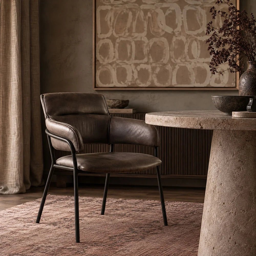 Whitcomb faux leather dining chair with a dark cushioned seat, padded back and curved arms stands beside a round table on a patterned dining room rug.