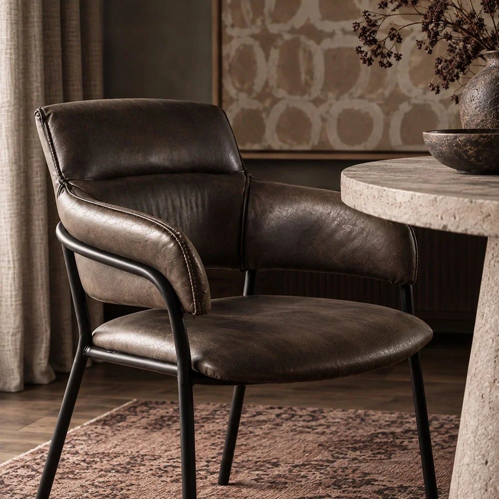 Whitcomb faux leather dining chair beside a pale round table, with dark brown upholstery, curved padded arms and slim black legs in a dining room.