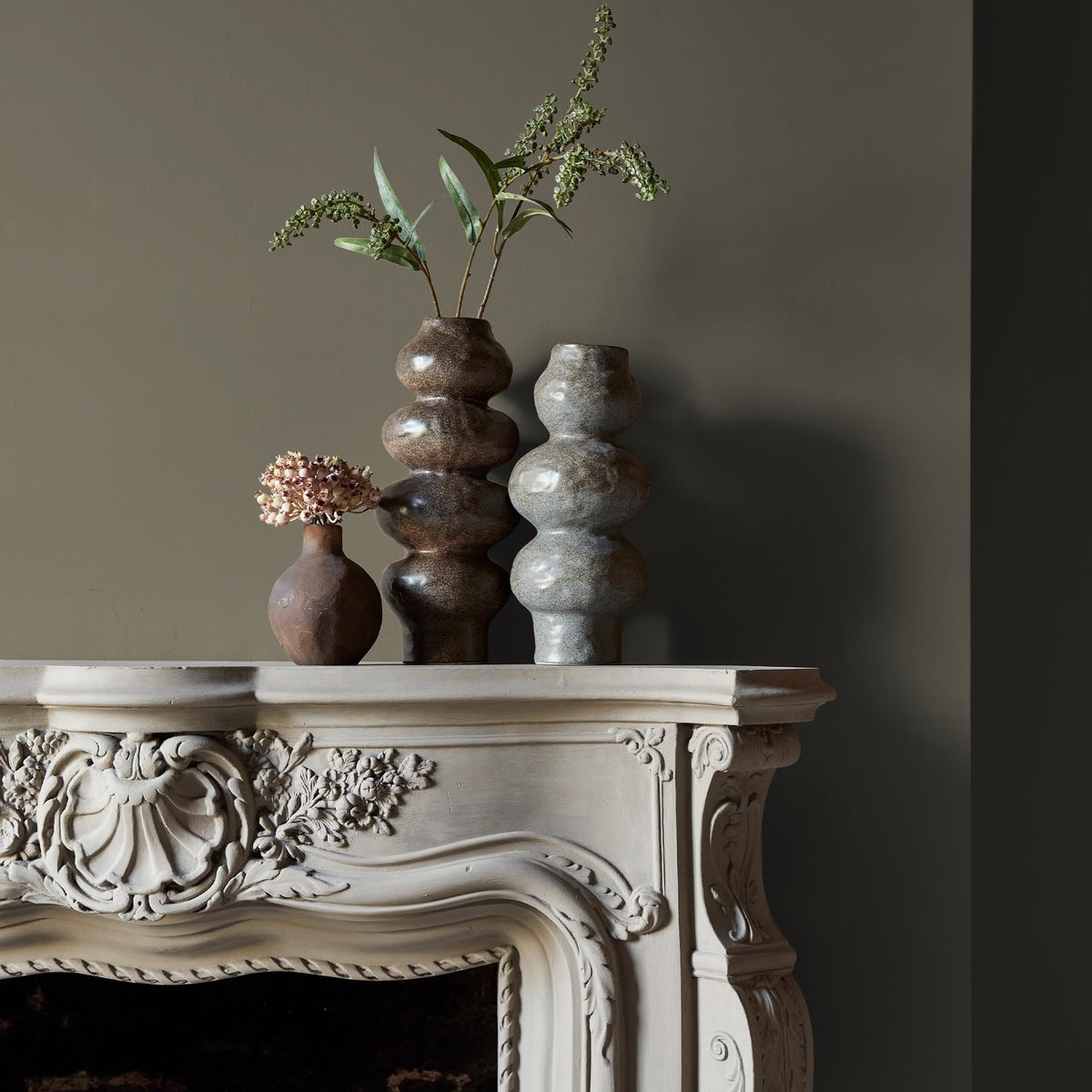 Three ceramic vases sitting on an intricate fireplace mantle in front of a wall in a sage green paint colour.