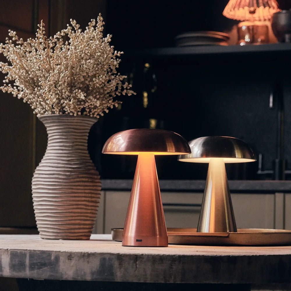 Zenith rose gold portable table lamp glows beside a silver lamp and ribbed vase, with a domed shade and tapered base on a console.