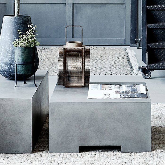 The Beaumont Coffee Table, a grey concrete square coffee table with a cutout leg design, on a sunny patio with a lantern.
