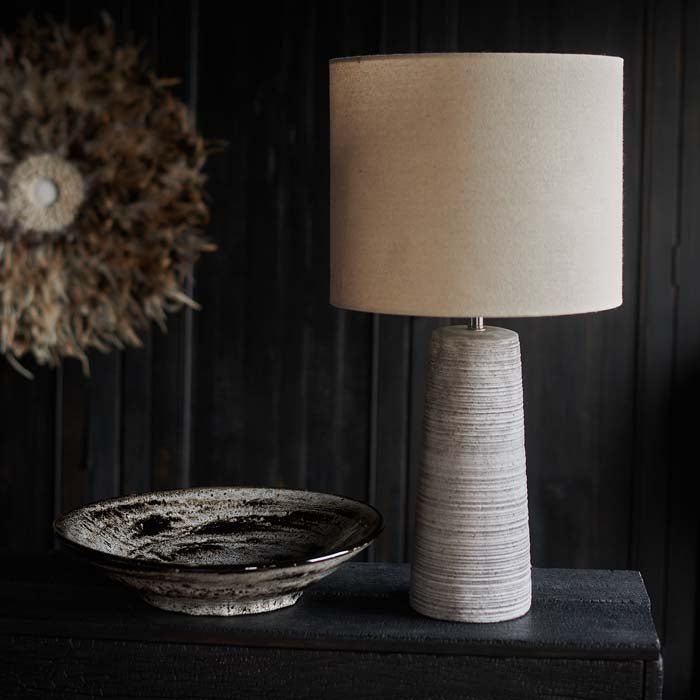 The Fria Table Lamp, with cream drum shade and grey textured base, against a plack wall and a feathered wall hanging.
