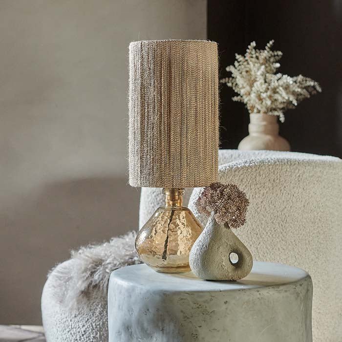 Styled image of a table lamp with a textured, pale orange glass base. The round glass lamp base is complimented by a tall lamp shade made of jute.