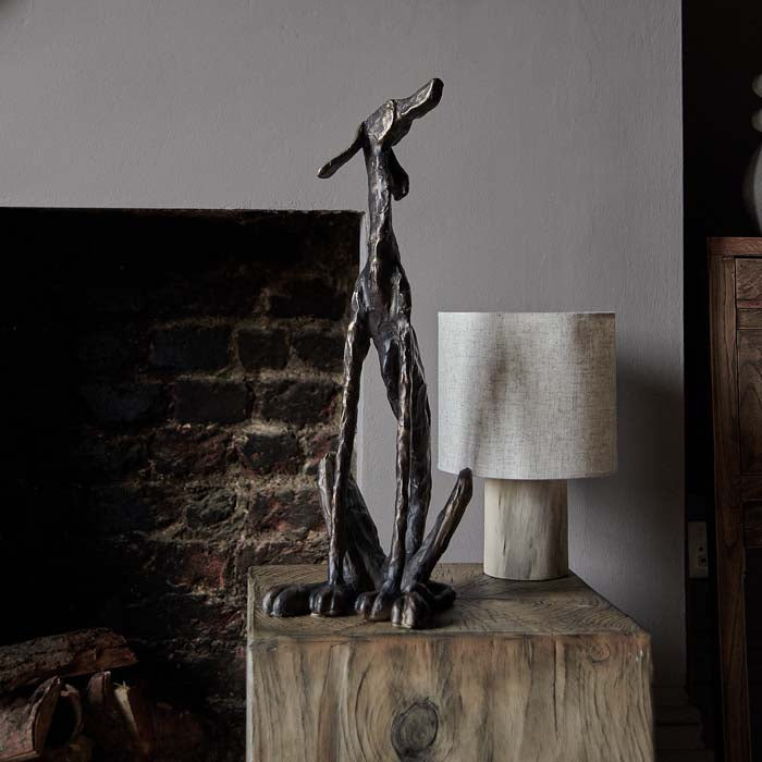 A bronze-look dog statue with an iron-look finish, placed on a wooden surface next to a lamp, with a brick wall and fireplace in the background.