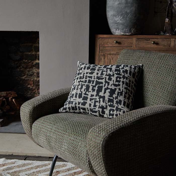 Square shaped woven cushion in natural and black tones in an abstract pattern on a green armchair.