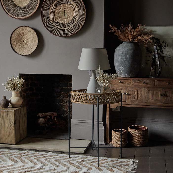 The Marino Rattan Side Table, a round rattan tray table with a lamp in front of a fireplace in a room with a boho vibe.