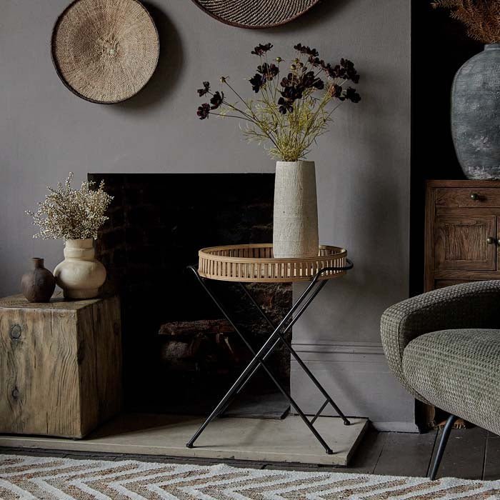 The Rosia Bamboo Side Table, a round slatted bamboo table with a black iron frame, next to a fireplace.