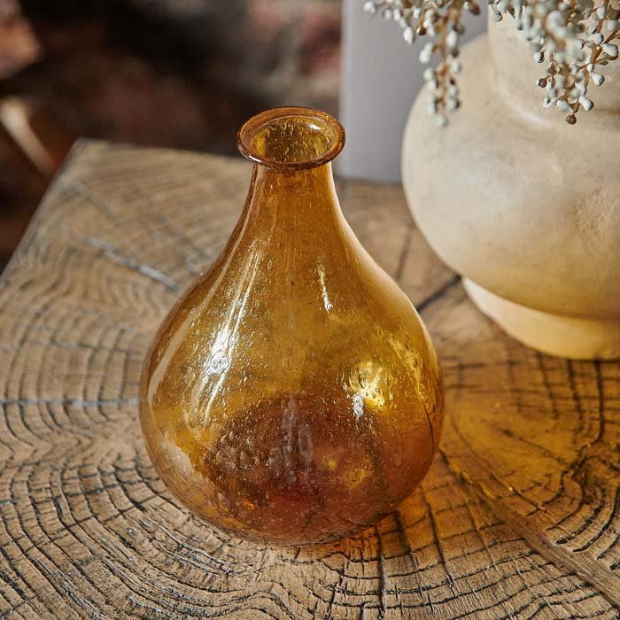 Amber glass bottle vase on wooden side table in an organic teardrop shape.
