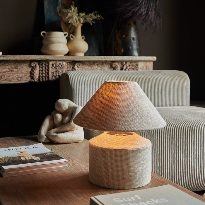 Warm light glows from a cream fabric table lamp. Styled on a coffee table. Luxury lighting and unique lighting styles from Abigail Ahern. 