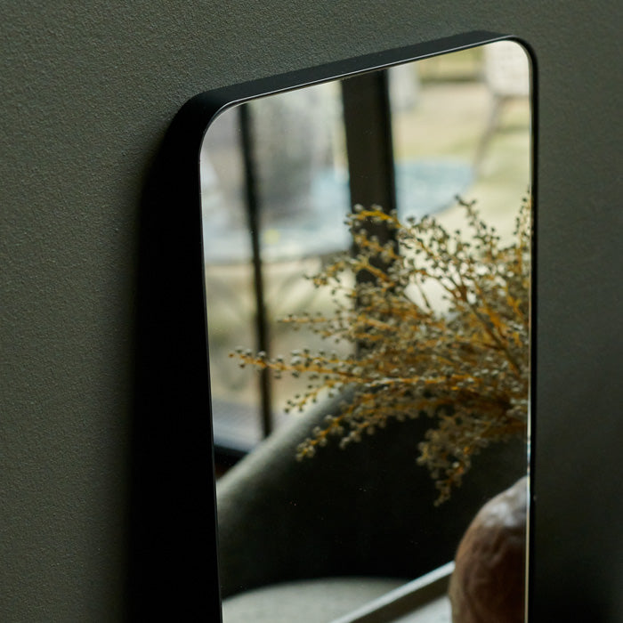Curved edge mirror in a thin black rectangular frame leaning against a green wall