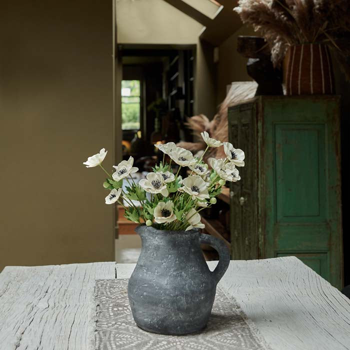 Rustic grey stone jug, on a kitchen table, filled with artificial flowers.