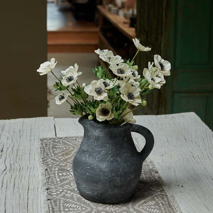 Artificial cream anemone flowers in stoneware jug, on wooden dining table.