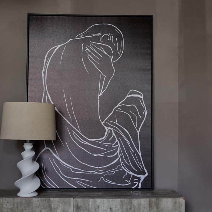 Large black framed figurative print leaning against a wall with a lamp in front
