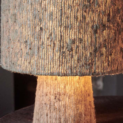 A close up of a textured fabric table lamp with a sort fabric base and a fabric drum shade in a neutral colour.