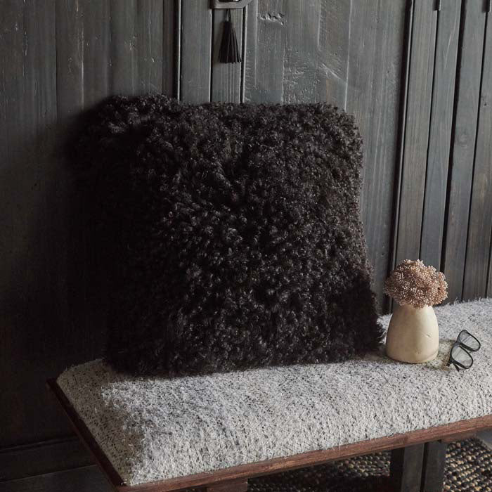 Shaggy black sheepskin cushion in a square shape displayed on upholstered wooden bench.