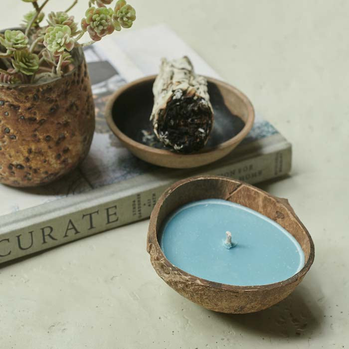 Blue coloured scented candle with a coconut shell holder sat in front of a burning smudge stick