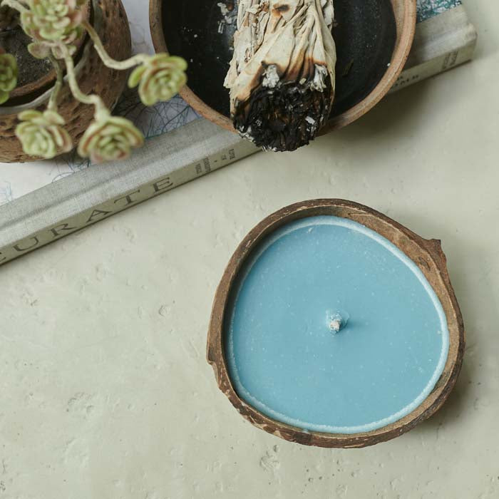 Blue coloured scented candle sat in a coconut shell on an off white table
