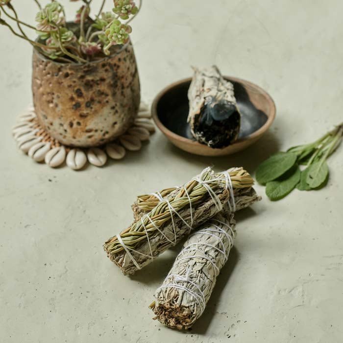 Three white sage smudge sticks with sweetgrass braided on top lying next to a burning smudge stick