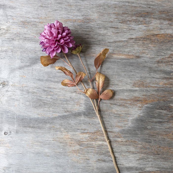 Artificial dahlia flower stem, with pinky-purple flower head and dried-look brown leaves.