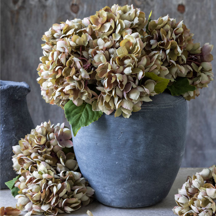 Grey stoneware vase filled with artificial hydrangea flowers in soft pinky-green tones.