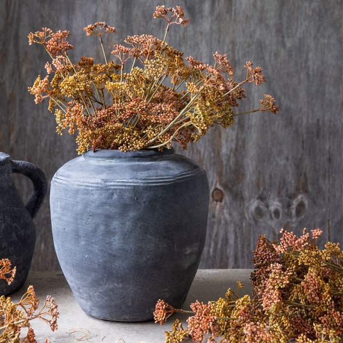 Grey stoneware vase filled with artificial meadow foliage in autumnal orange tones.