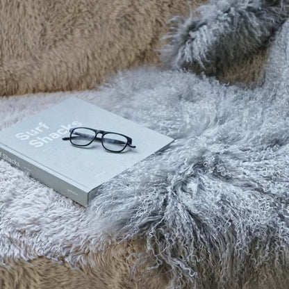 Closeup image of a long haired sheepskin in a pale grey colour with a book and some glass sitting on it