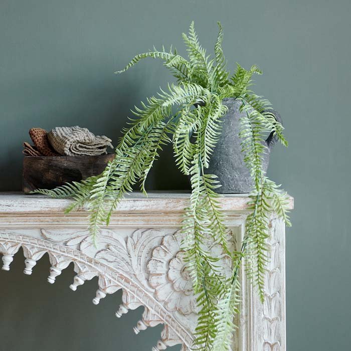 Green fake ferns in a grey vase sitting on top of a carved console.