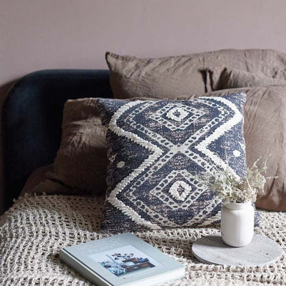 Square decorative cushion with monochromatic print and textured embroidery. The pillow is in front of several other cushions on the bed, next to a faux plant and hardback book.