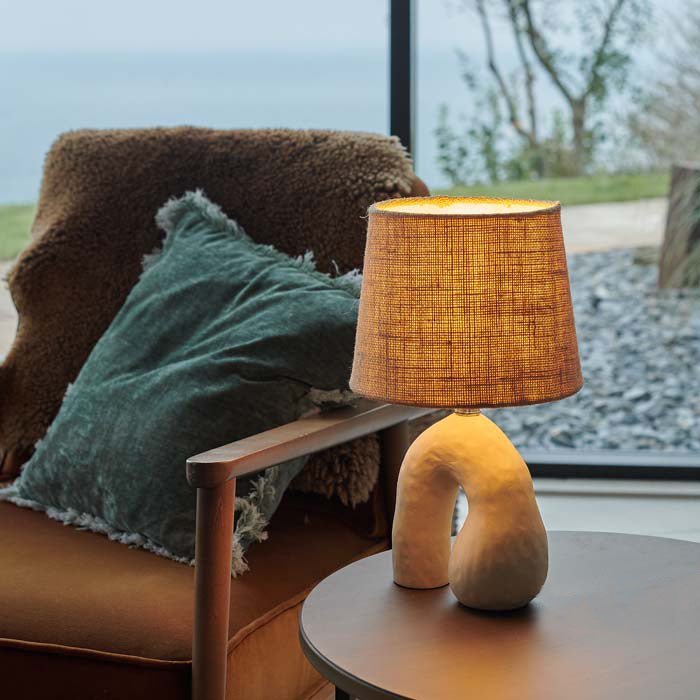 Styled image of a sculptural, U-shaped cream ceramic lamp with a woven shade. This ceramic table lamp gives off a lovely, cosy glow.