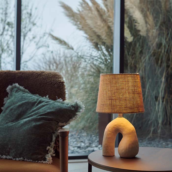 Styled image of a cream, organic shaped ceramic lamp with a woven shade, giving off a soft glow.