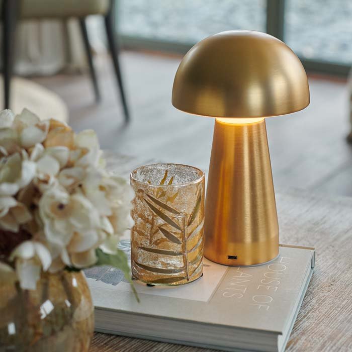 A gold metal curvaceaous mushroom shaped wireless and portable lamp by Abigail Ahern, known for her rule breaking designs and stylish interiors. Styled on a coffee table with some books and some faux flowers, this small but mighty curvy lamp is super flexible and dimmable too, pop all over the house, style next to your bath, bedside. 