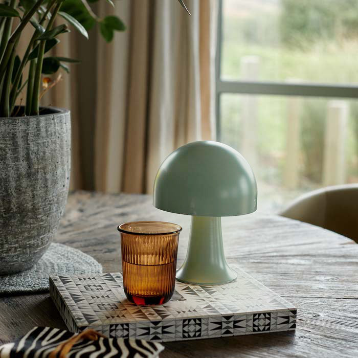 Small portable LED table lamp in forest green. Styled on a coffee table, book and drinking glasses in a stylish interior. These dome shaped lamps are battery operated, therefore no need for plug sockets. These small curvy lamps are super flexible to pop all over. the house, style next to your bath, bedside or create intrigue to your tablescapes. Available in another colourway as well as this soft timeless green colour. 
