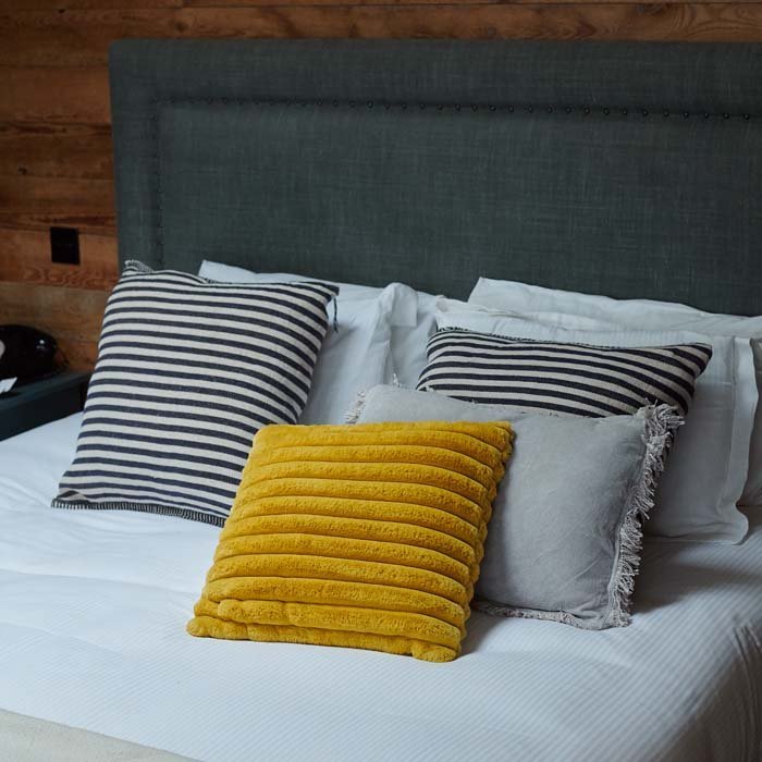 The Kinsley Cushion, a square cushion in vibrant mustard coloured faux fur with ridged detail, with cushions on a bed.