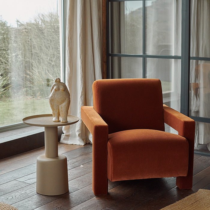 Abigail Ahern Phoenix Velvet Armchair in burnt orange velvet, showcasing its softly padded seat and backrest, distinctive angular armrests and relaxed reclined shape within a sophisticated modern interior.