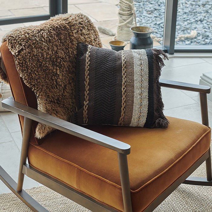 The Piper Cushion, a square cushion featuring stripes of varying widths in shades of brown and a dark brown fringe at one end.