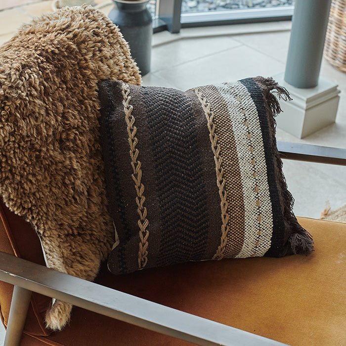 The Piper Cushion, a square cushion featuring stripes of varying widths in shades of brown and a dark brown fringe at one end.