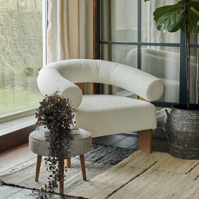 Abigail Ahern Ryton Cream Boucle Armchair in soft cream boucle upholstery, featuring a curving mid-century inspired silhouette with a rounded back and cylindrical armrest, styled beside a window with greenery and natural light.