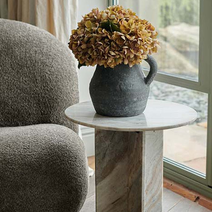 The Tortona Side Table, a round table in rich brown and grey hued marble with a cross shaped pedestal base.