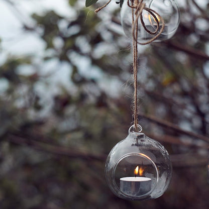 Detail image of a circular, glass hanging tea light holder with a lit tealight, hanging from a branch by twine