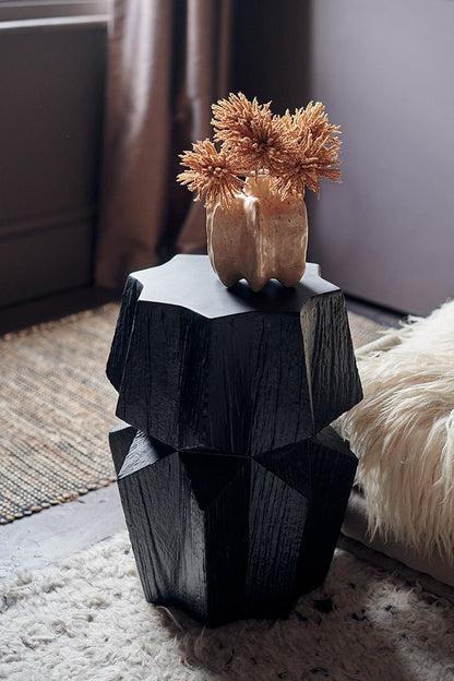 Styled image of a chunky, sculptural black side table with a wood style finish. The black side table has been styled with a vase with a faux bouquet of fake flowers.