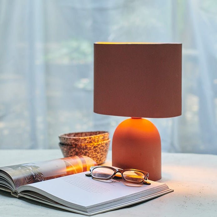 A browny pink lit table lamp with a matching shade next to a book and some glasses.
