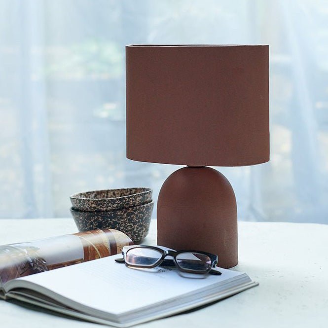 A terracotta coloured table lamp with a matching shade next to an open book and some glasses.