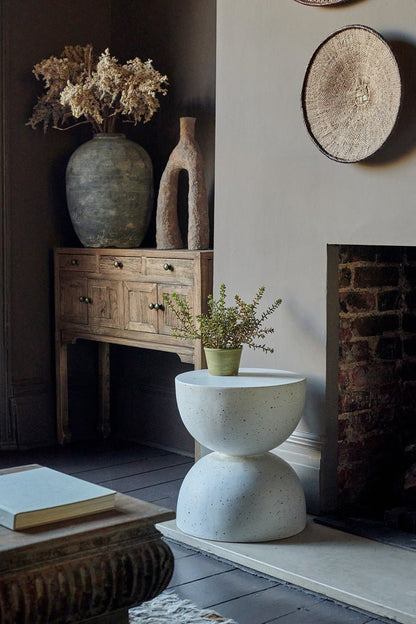 Styled image of an hourglass shaped white side table with a small faux plant on it. In the background is a console with some ceramic vases.