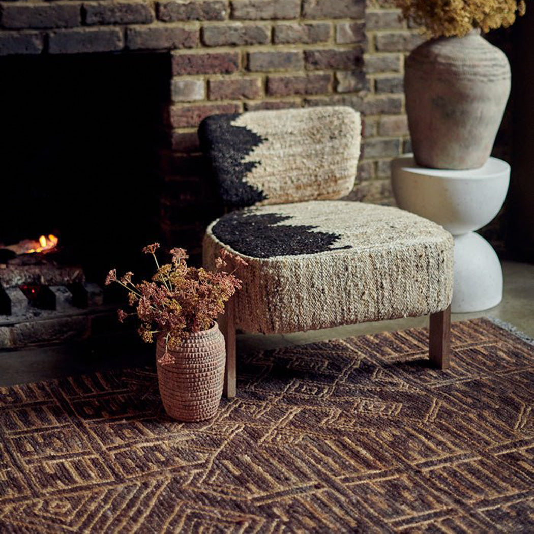 A boho accent chair sitting on a woven jute rug with a vase of artificial flowers next to it.