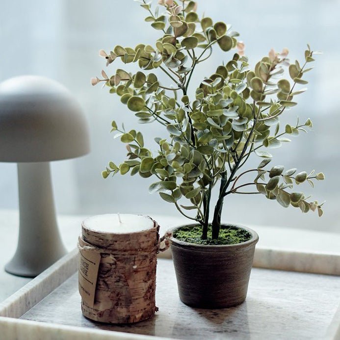 A small potted artificial plant with green leaves next to a candle.