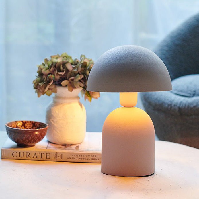 A small pale grey table lamp with a round base and a matching dome shade next to a small vase of faux flowers.